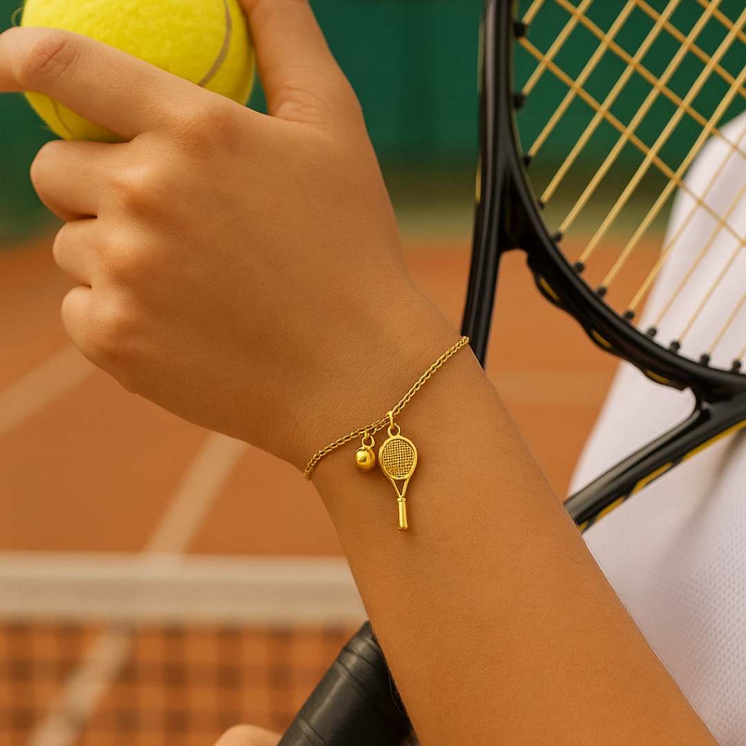 Gold bracelet with tennis racket and ball charm on a wrist against a beige background from House of Pehr