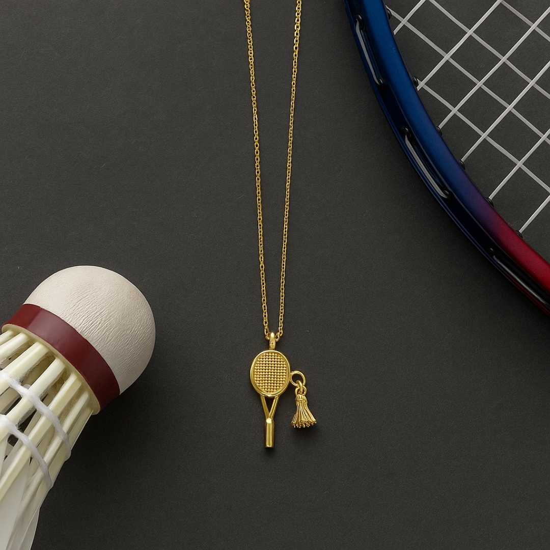 Gold necklace with a badminton racket pendant next to a badminton shuttlecock and racket on a dark surface from House of Pehr
