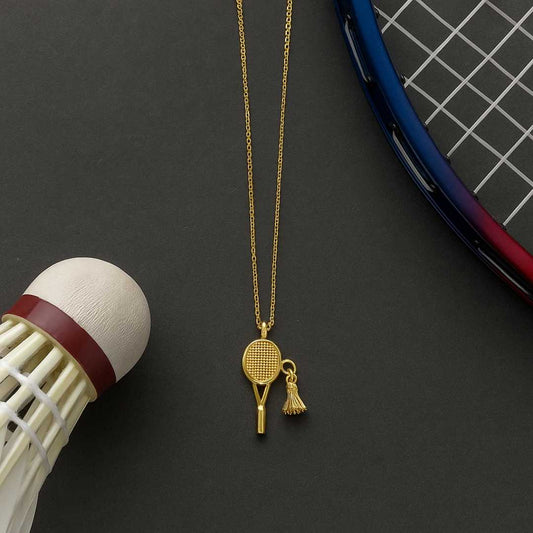 Gold necklace with a badminton racket pendant next to a badminton shuttlecock and racket on a dark surface from House of Pehr