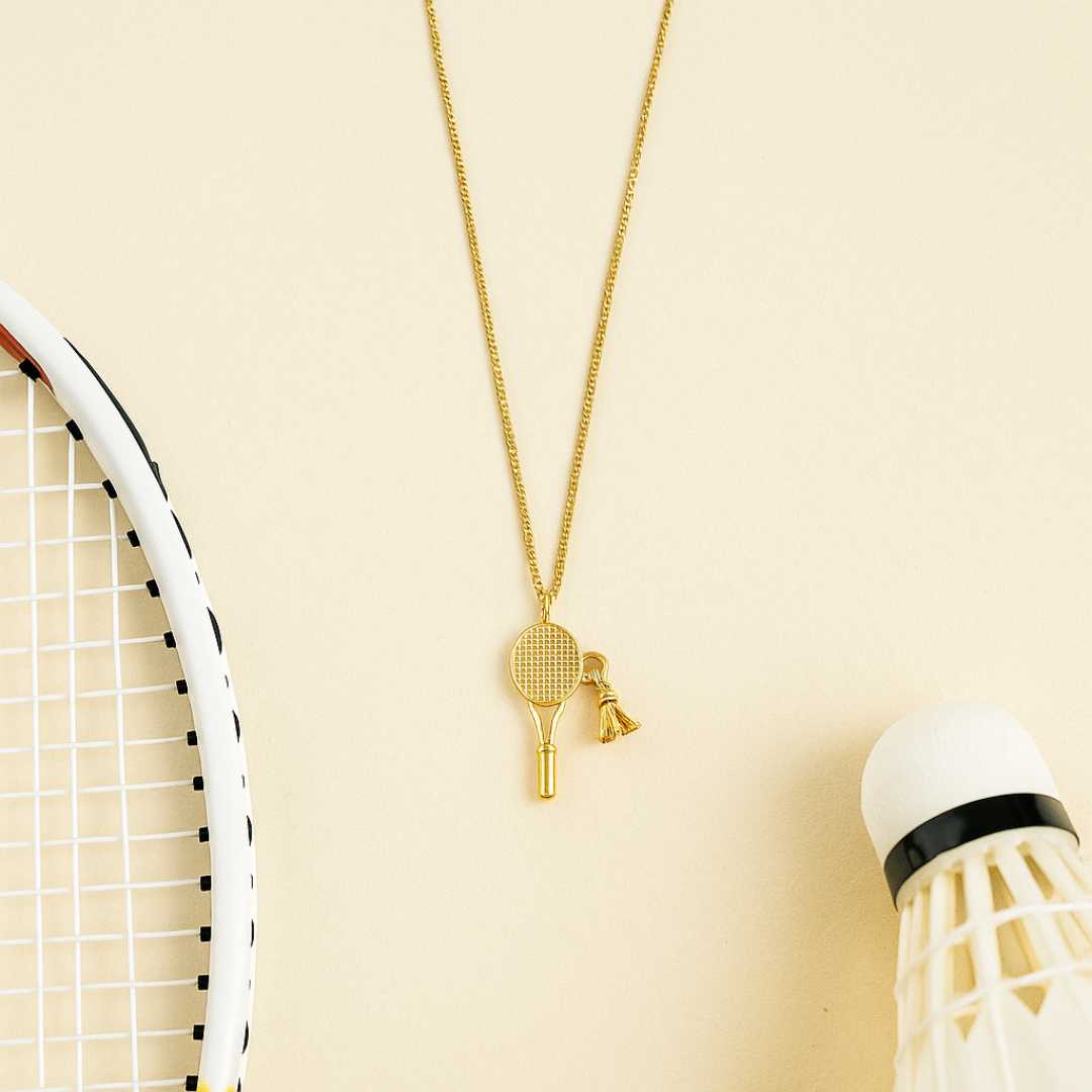Gold necklace with a badminton racket pendant next to a badminton shuttlecock and racket on a dark surface from House of Pehr