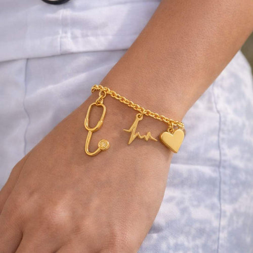 Gold bracelet with stethoscope, heart, and heartbeat design on a wrist against a white background from House Of Pehr 