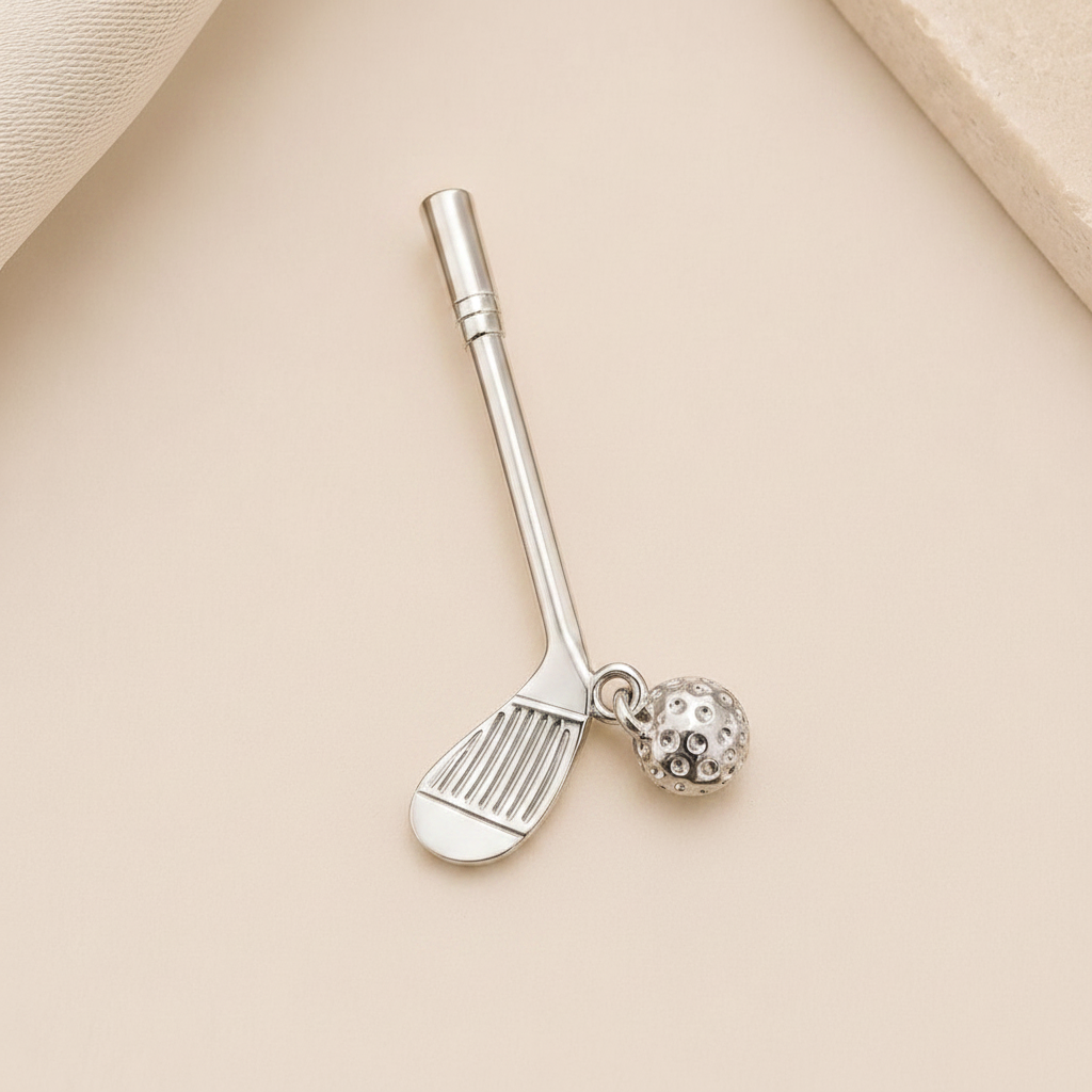 A silver-toned metal nail clacker shaped like a golf club, with a small dangling golf ball charm, displayed on a light beige textured background.