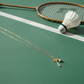 Gold necklace with a badminton racket charm on a green surface with a badminton racket and shuttlecock from house of pehr