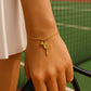 Gold bracelet with tennis racket and ball charm on a wrist against a beige background from House of Pehr