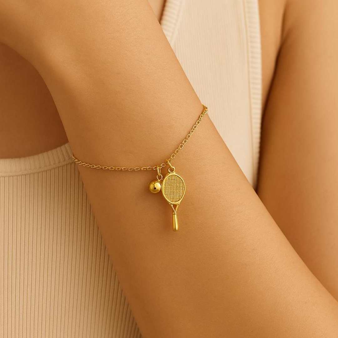 Gold bracelet with tennis racket and ball charm on a wrist against a beige background from House of Pehr